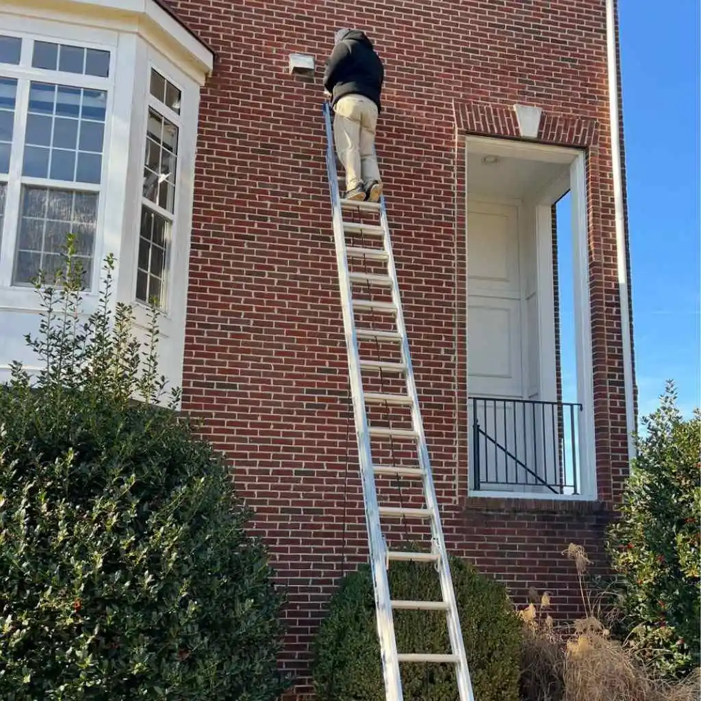 Technician on a tall ladder inspecting a high exterior vent to resolve allergy symptoms from air ducts caused by airflow blockages.